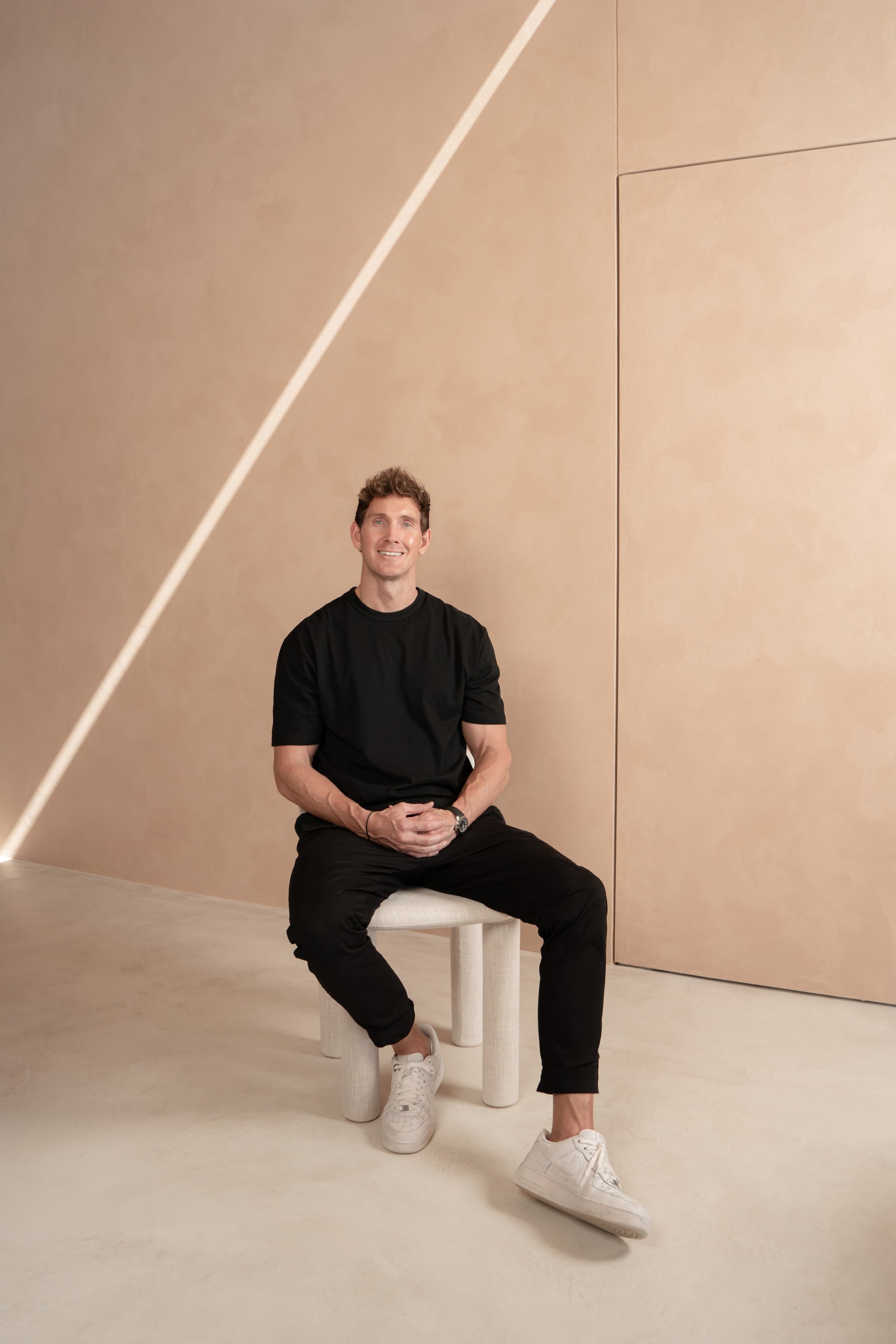 Founder seated on a cream stool against a warm-plaster wall with diagonal light line. Graya